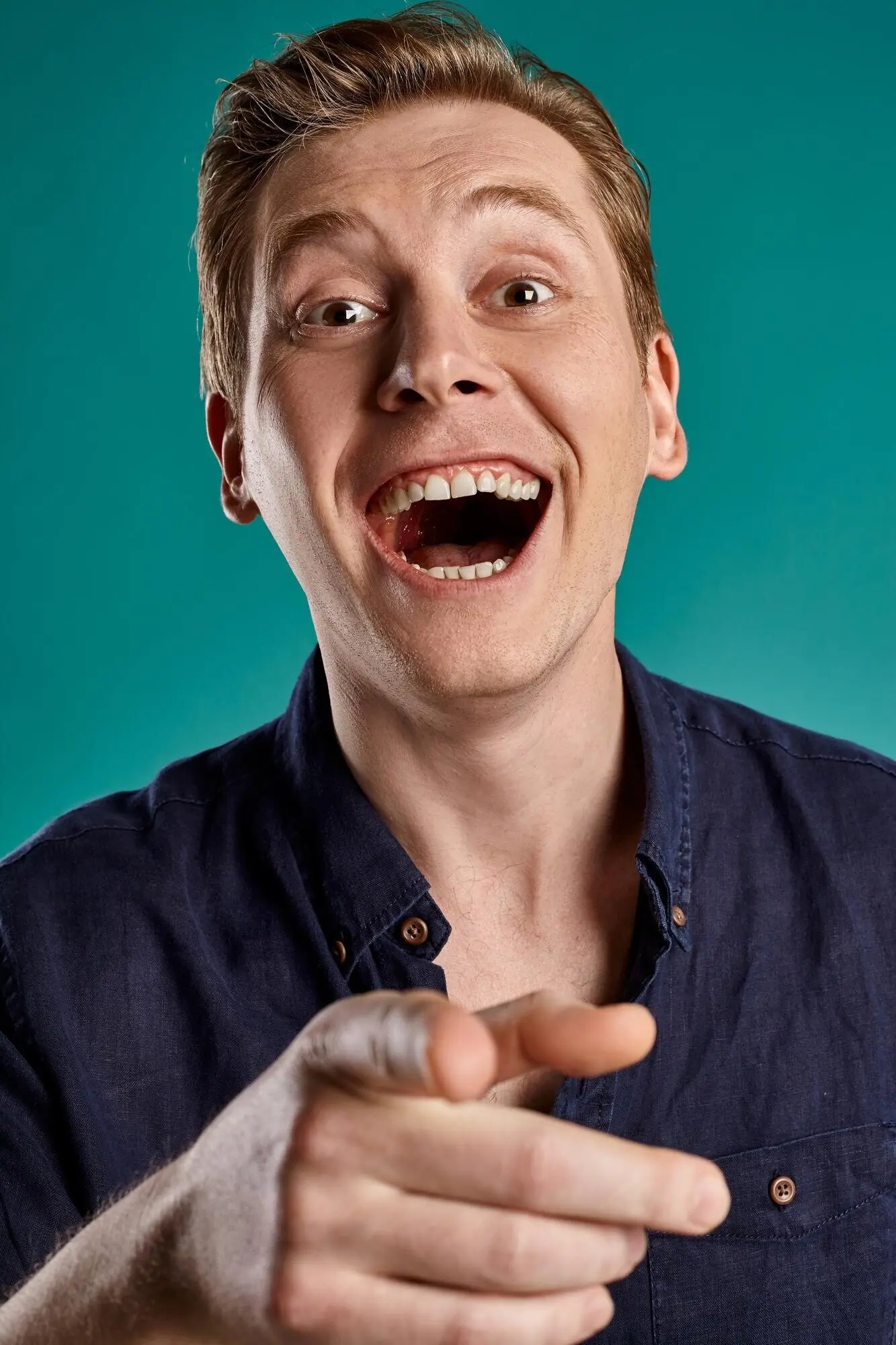 Nahes Porträt eines jungen, witzigen Rothaarigen in einem stylischen marineblauen T‑Shirt, der in die Kamera schaut und dabei jemanden auslacht, während er vor einem blauen Studiohintergrund posiert. Menschliche Gesichtsausdrücke. Aufrichtige Emotionen.